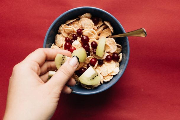 mão feminina adicionando kiwi para tigela com flocos de milho crocante e cranberries no fundo vermelho, café da manhã saudável
 - Foto, Imagem