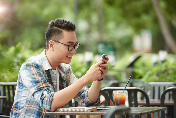 Glimlachend Aziatische man in glazen lezen van berichten in zijn smartphone - Foto, afbeelding