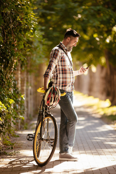 Красивый бизнесмен едет работать на велосипеде. Он стоит рядом с велосипедом и использует смартфон
. - Фото, изображение