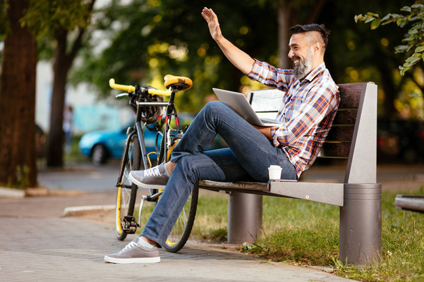 Usmívající se neformální podnikatel mává Hello na přestávku na kávu. On sedí na lavičce a práci na notebooku, vedle lavičky spočívá na kole. - Fotografie, Obrázek