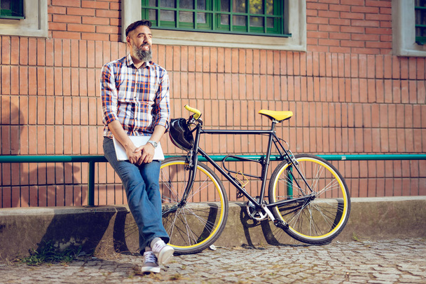 Χαμογελώντας casual επιχειρηματία έχοντας ένα διάλειμμα. Στέκεται μπροστά από το κτίριο δίπλα στο ποδήλατο και κρατώντας το laptop. - Φωτογραφία, εικόνα