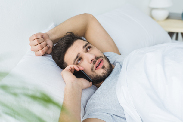 bearded man lying on bed and talking on smartphone in the morning - Фото, изображение