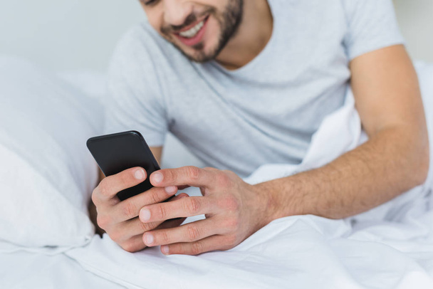 smiling bearded man lying on bed and using smartphone - Photo, Image