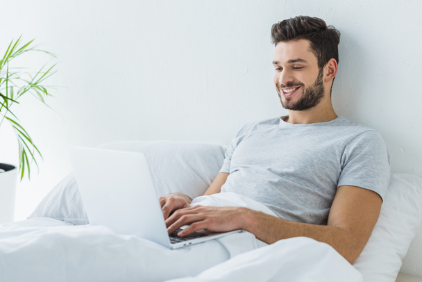 smiling man lying on bed and using laptop - Фото, изображение