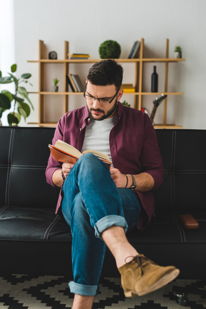 Young male in glasses sitting on leather couch and reading book - Photo, image