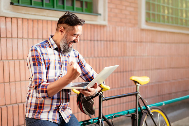 Un homme d'affaires souriant qui travaille sur un ordinateur portable. Il se tient devant le bâtiment à côté du vélo
. - Photo, image