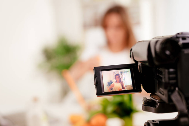 Mulher bonita filmando seu blog transmitido sobre comida saudável em casa. Foco selectivo. Concentre-se em primeiro plano, na câmera
. - Foto, Imagem