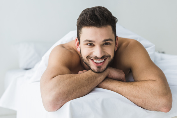 schöner, fröhlicher Mann liegt auf weißem Bett und schaut in die Kamera - Foto, Bild