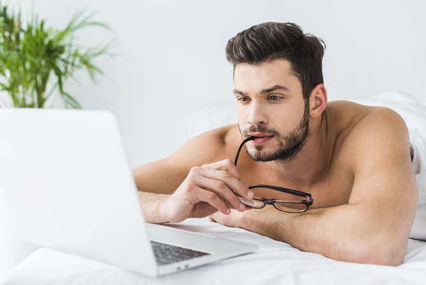 homem pensativo com óculos usando laptop na cama pela manhã
 - Foto, Imagem