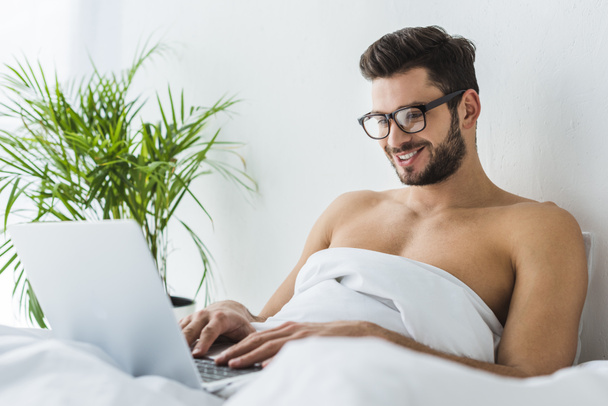 handsome smiling man using laptop in bed - 写真・画像