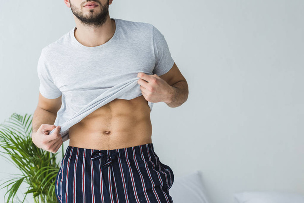 cropped view of sexy man taking off his t-shirt in bedroom - Φωτογραφία, εικόνα
