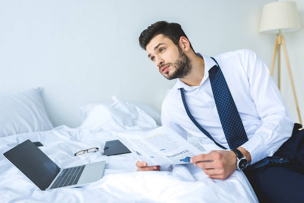 thoughtful businessman working with newspaper and laptop on bed in morning - Foto, Imagem