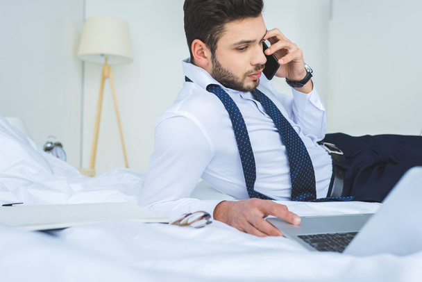 knappe zakenman liggend op bed tijdens het werken met de laptop en smartphone - Foto, afbeelding