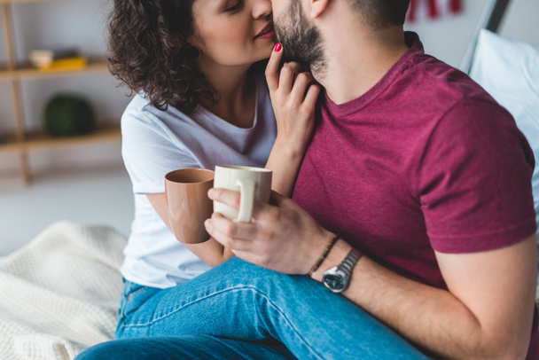 Recortado vista de sonriente mujer besando novio mientras él sosteniendo tazas
 - Foto, imagen