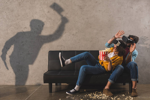 Ombra di uomo tenendo ascia e coppia spaventata con popcorn utilizzando cuffie realtà virtuale
 - Foto, immagini