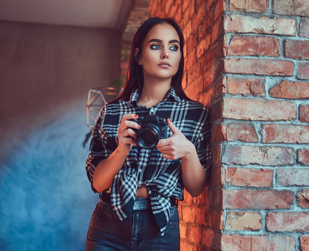 Portrait of an attractive brunette girl dressed in a flannel shirt and jeans with camera leaning on the wall in a room with loft interior. - Foto, Imagem