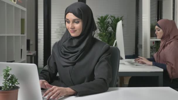 Young beautiful girl in black hijab typing on laptop, looking at camera and talking, speaking. 60 fps - Video