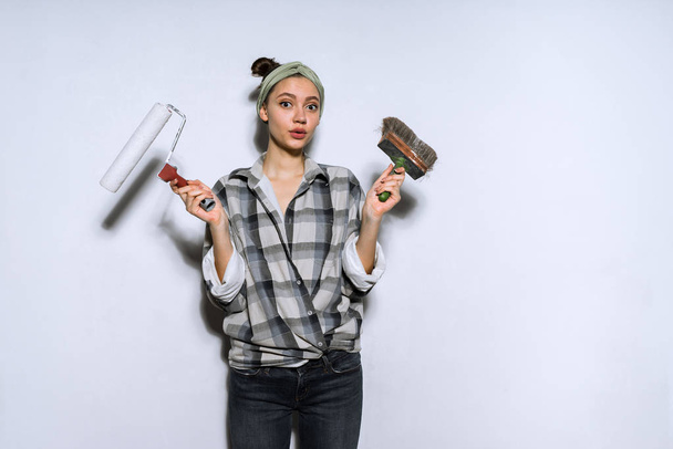 a young girl in a plaid shirt doing repairs in her new apartment, holding a roller and a painting for walls - Fotoğraf, Görsel