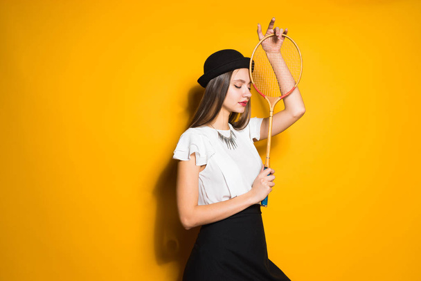 young attractive model girl in fashionable black hat posing on yellow background, holding a racket for playing tennis - Photo, image