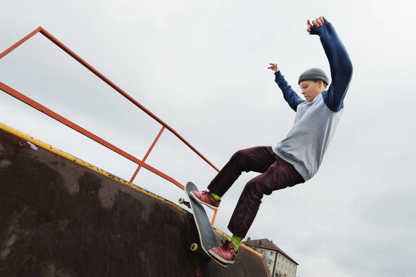 A teenager skateboarder in a hat does a Rocks trick on a ramp in a skate park against a cloudy sky and sleeping area. The concept of urban style in sport - Foto, immagini