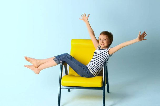 Cute Young Girl, Wearing blue Jeans And  Striped T-Shirt, Sitting in Yellow Arm-Chair Over Blue Background and Holding Hands Up. - Фото, изображение