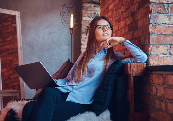 Portrait of charming brunette businesswoman in glasses and blue shirt working online with laptop and sitting on a sofa in a room with loft design. - Fotoğraf, Görsel