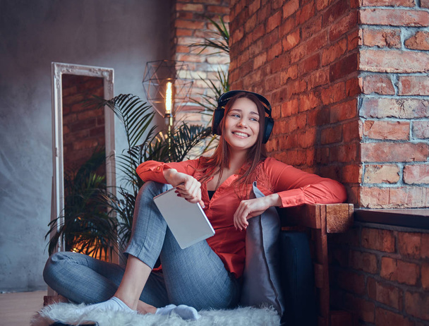 Een aantrekkelijke lachende brunette meisje gekleed in casual kleding met behulp van een tablet terwijl u luistert naar de spannende muziek via goede hoofdtelefoons. Ontspannen in een kamer met loft interieur. - Foto, afbeelding