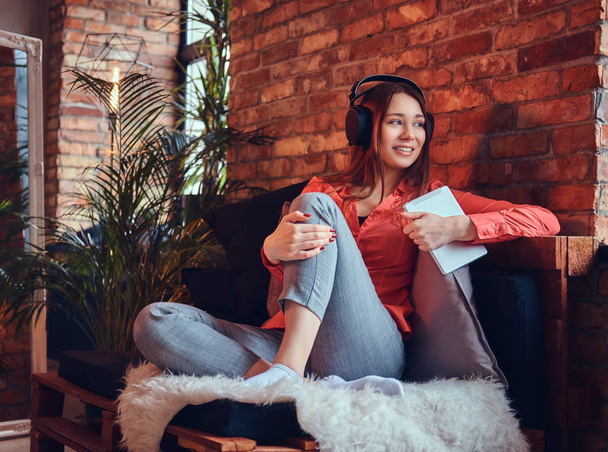 Atrakcyjny brunetka uśmiechający się ubrany w ubranie przy użyciu tabletu podczas słuchania ekscytujące muzyki poprzez dobre słuchawki. Relaks w pokoju z wnętrza loft. - Zdjęcie, obraz