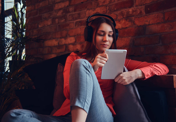 Een aantrekkelijke hipster meisje gekleed in casual kleding glimlachen terwijl u luistert naar de spannende muziek via goede hoofdtelefoons. Ontspannen in een kamer met loft interieur. - Foto, afbeelding