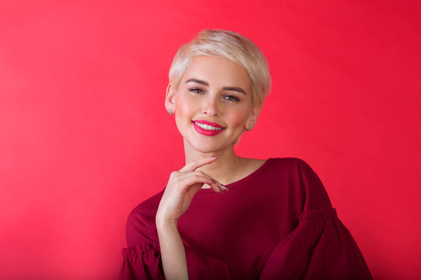 beautiful young girl with snow-white smile and healthy teeth with short hairdo on red background - Photo, image