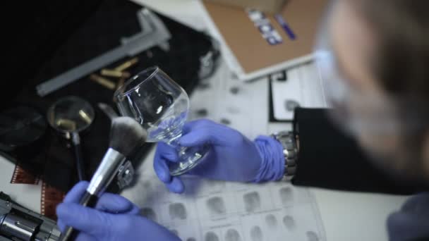 Etsivä hanskoissa nostamassa sormenjälkiä viinilasista, käyttäen harjaa ja jauhetta
 - Materiaali, video