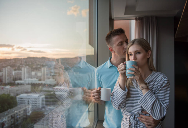 romantic happy young couple enjoying evening coffee and beautiful sunset landscape of the city while standing by the window - Foto, imagen