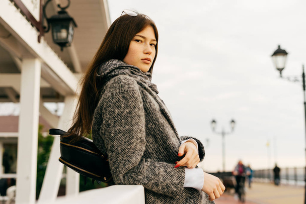 stylish urban girl with dark hair, in a gray coat walking through the streets of the city and posing - 写真・画像