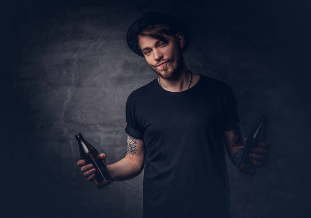 Portrait of an attractive bearded male with tattooed arms, dressed in a black t-shirt and hat holds bottled beer. - Fotó, kép