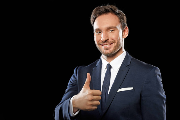 Handsome smart businessman in a classic office suit and tie showing an okay sign with thumbs up isolated on a black background, a guy you can trust - Foto, afbeelding