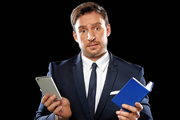 Hezký chytrý podnikatel v klasické úřadu obleku a kravatě drží notebook a telefon ukazuje tvrdou práci. izolované na černém pozadí - Fotografie, Obrázek