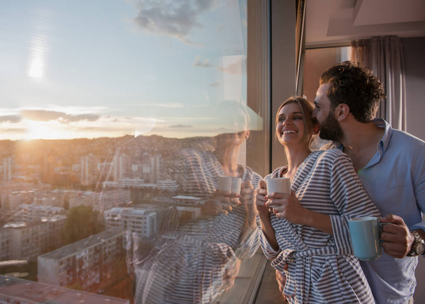 romantic happy young couple enjoying evening coffee and beautiful sunset landscape of the city while standing by the window - Valokuva, kuva