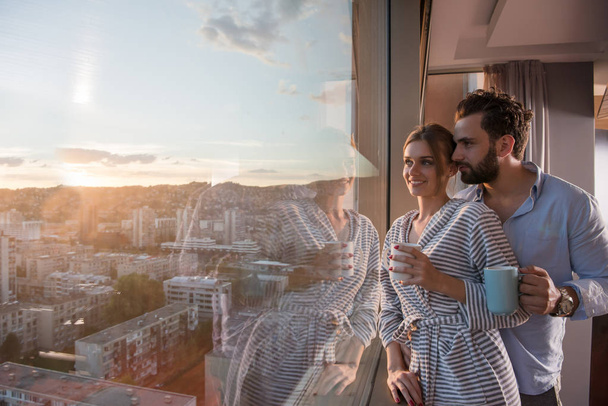 romantic happy young couple enjoying evening coffee and beautiful sunset landscape of the city while standing by the window - Fotó, kép