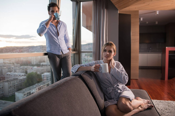 romantic happy young couple enjoying evening coffee and beautiful sunset landscape of the city while standing by the window - Foto, Imagen