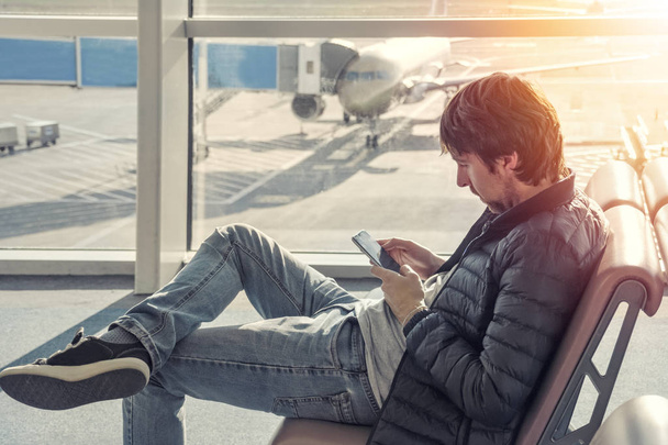 Молодой кавказский мужчина в джинсах и куртке сидит в зале ожидания аэропорта, используя мобильный телефон. Онлайн дистанционное банковское обслуживание, бронирование отелей, мобильность, чтение новостей с помощью 4g, 3g
. - Фото, изображение