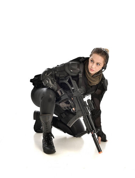 Pełna długość Portret Kobieta w czarnej zbroi taktyczne, Przyczajony poza i trzyma broń, na białym tle na tle białego studia. - Zdjęcie, obraz