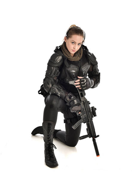 retrato de comprimento total da mulher vestindo armadura tática preta, postura agachada e segurando uma arma, isolado no fundo do estúdio branco
. - Foto, Imagem