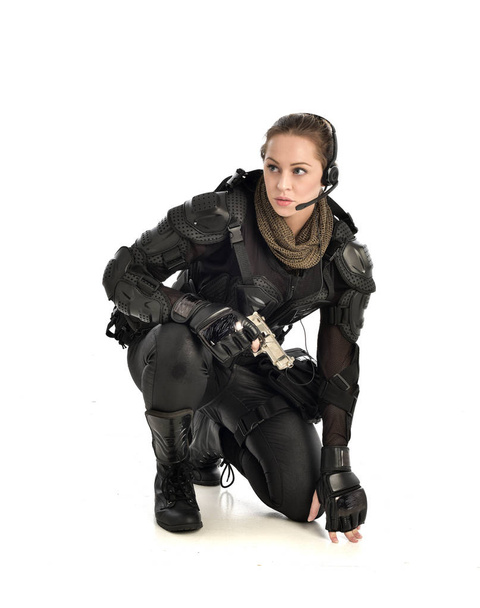 女性の黒の戦術的な鎧、しゃがみポーズを身に着けている白いスタジオ背景に分離した武器を持ちの完全な長さの肖像画. - 写真・画像