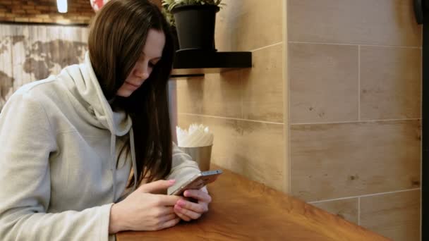 Pensive young brunette woman in a bright shirt using internet in her cellphone sitting in cafe. - Metraje, vídeo