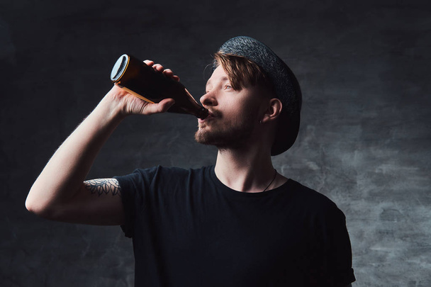 Retrato de un atractivo hombre hipster con brazos tatuados, vestido con una camiseta negra y sombrero bebe cerveza embotellada
. - Foto, Imagen