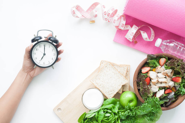 Salad bowl, whole wheat bread, milk, measuring-tape, yoga mat and woman's hand holding alarm clock, Healthy diet concept - Photo, Image