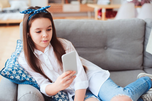 happy preteen girl relaxing at home on cozy couch and listening music, using smartphone - 写真・画像