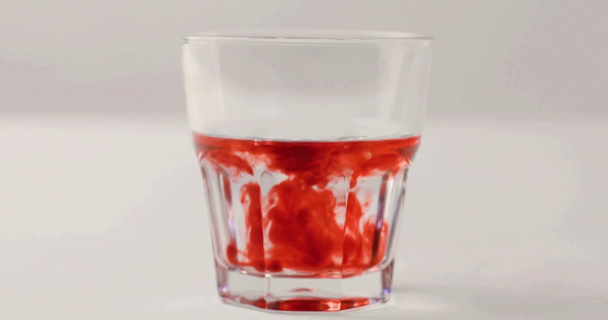 Remolinos de jarabe rojo en vaso con agua sobre fondo blanco
 - Metraje, vídeo