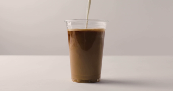 Ρίχνει γάλα σε πλαστικό ποτήρι με καφέ σε λευκό φόντο με αντίστροφη πλάνα - Πλάνα, βίντεο