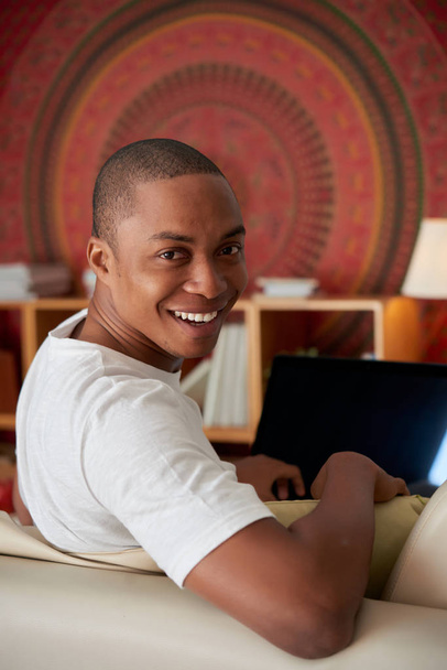 Портрет веселого молодого чернокожего человека, сидящего на диване и поворачивающегося к камере
 - Фото, изображение